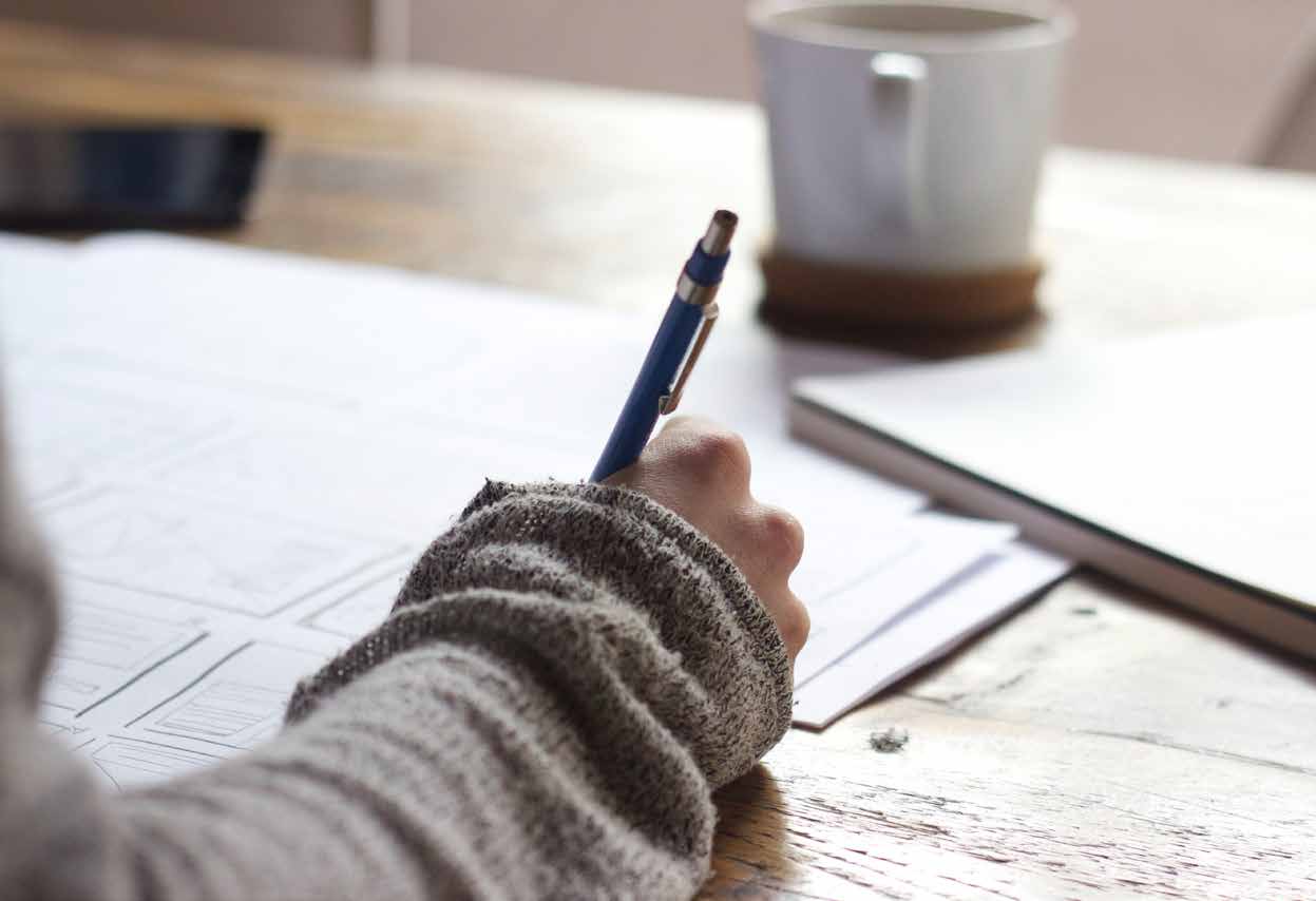 A hand writing notes on paper beside a cup of coffee